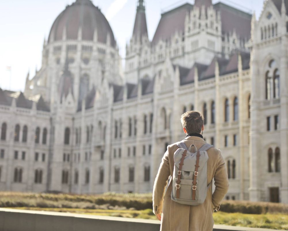 male-wearing-brown-coat-backpack-near-hungarian-parliament-building-budapest-hungary(1)